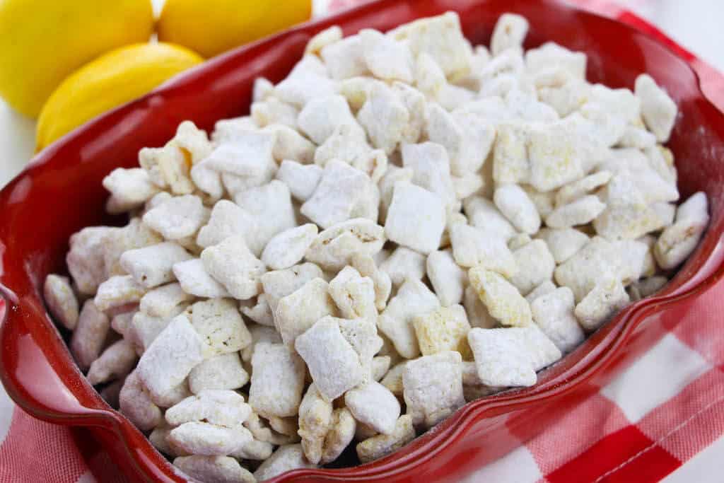 close up of lemon flavored muddy buddies in a red bowl with lemons next to it. 